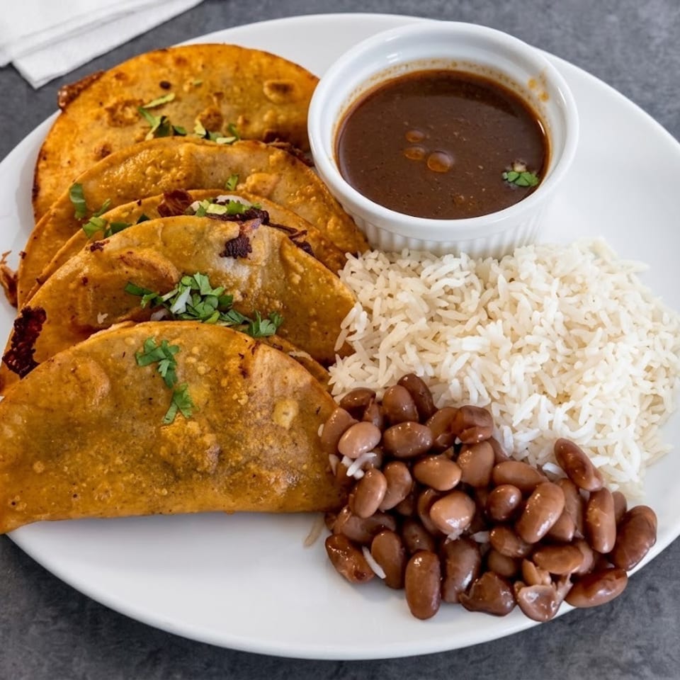 Plate of crispy tacos garnished with cilantro, served with white rice, pinto beans, and a cup of brown dipping sauce.
