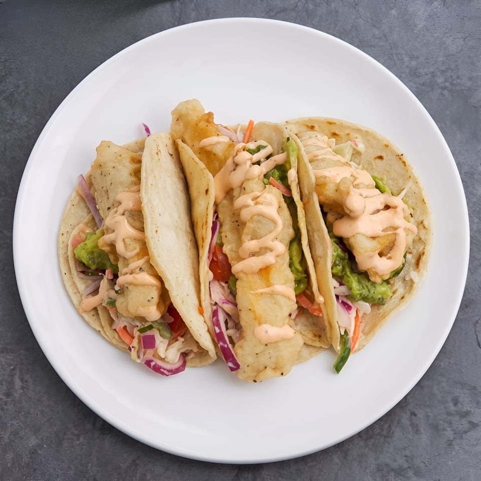 Three fish tacos topped with creamy sauce, guacamole, shredded cabbage, carrots, and red onion on a white plate.
